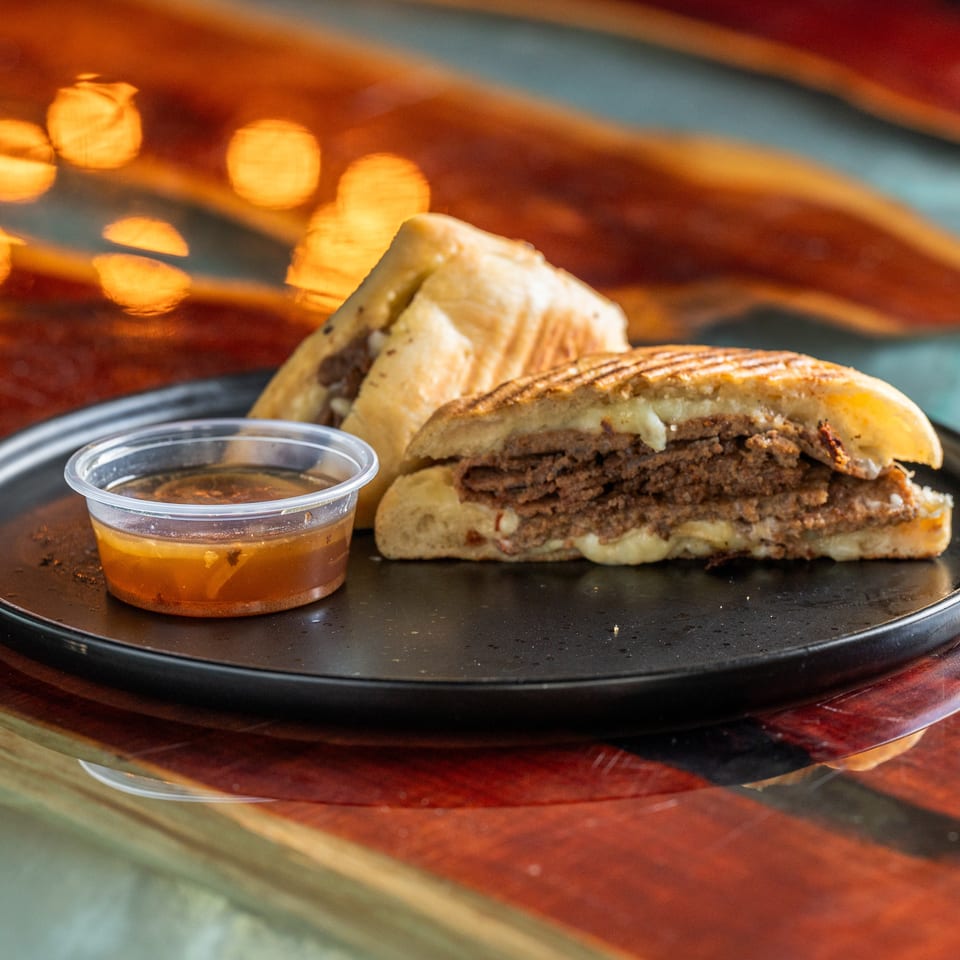 Double-patty cheeseburger on a black plate with a grilled sandwich behind it and a cup of dipping sauce.