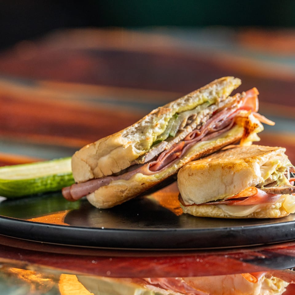 Two toasted deli sandwiches layered with meats, cheese, lettuce, and tomato on a black plate with pickle slices.
