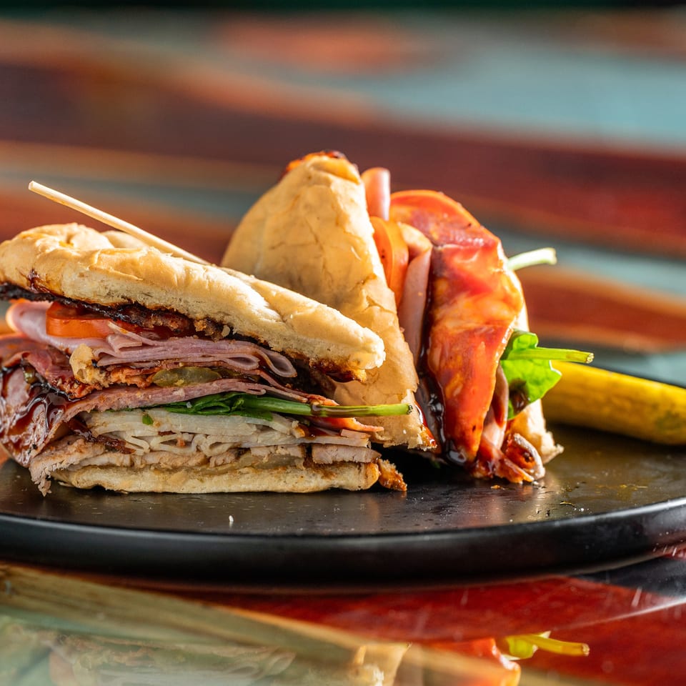 Stacked deli sandwich with sliced meats, cheese, vegetables, and sauce served alongside pickles on a black plate.