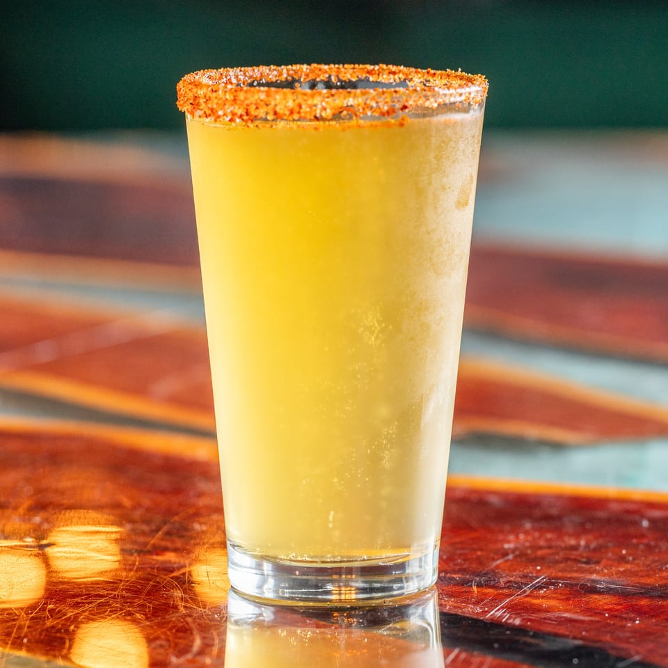 Yellow cocktail in a tall glass with a chili-salt rim, displayed on a reflective wooden bar.