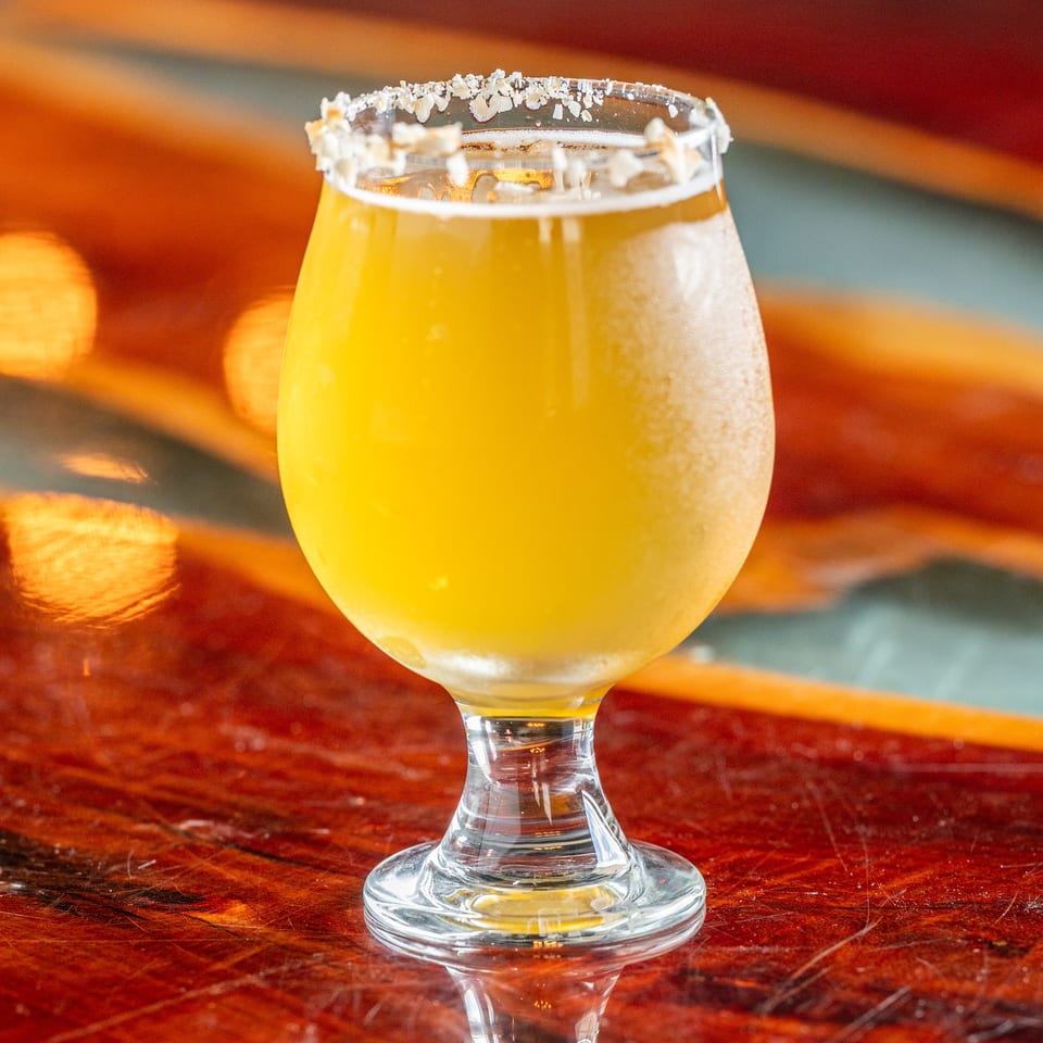 Golden cocktail in a stemmed glass with a coarse salt rim, displayed on a polished wooden bar.