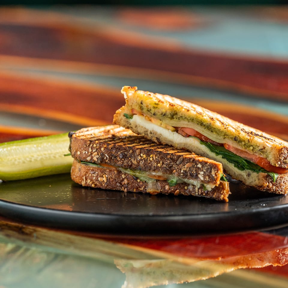 Grilled vegetable sandwich on seeded bread with pesto, tomato, greens, and a pickle spear on a black plate.