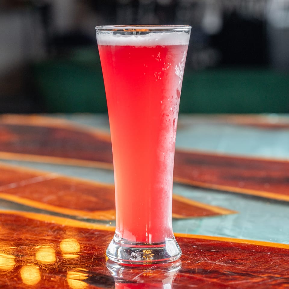 Bright red frothy drink served in a tall glass on a polished wooden restaurant table.