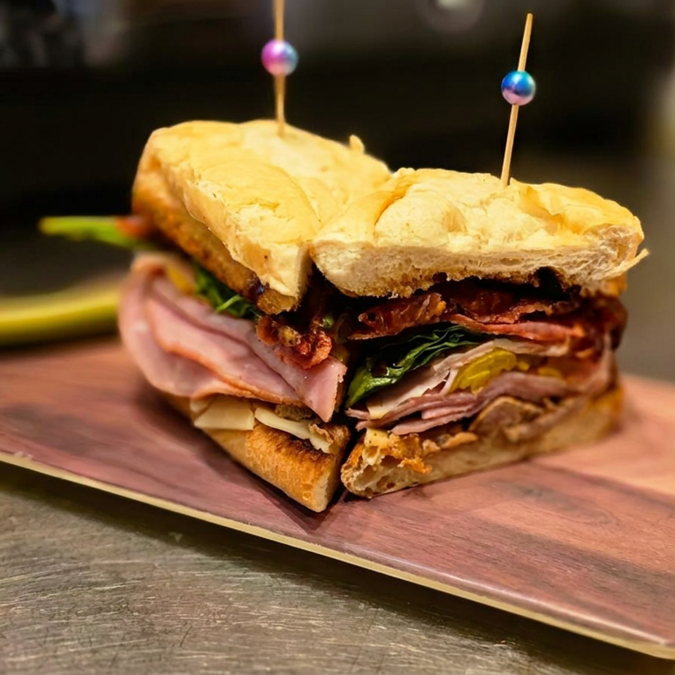 Layered deli sandwich with ham, vegetables, cheese, and crispy bacon served on a wooden board with decorative picks.